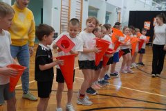 Na zdjęciu widać grupę Dzieci stojących w rzędzie w sali gimnastycznej. Trzymają one duże pomarańczowe pachołki i biorą udział w ćwiczeniu ruchowym, polegającym na przekładaniu piłeczki z pachołka do pachołka bez użycia rąk. Obok Uczniów stoją Nauczyciele, którzy nadzorują aktywność.