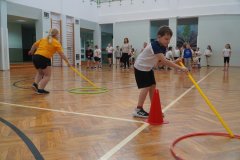 Na zdjęciu widać Uczniów uczestniczących w zajęciach ruchowych w sali gimnastycznej. Na pierwszym planie dwoje Dzieci wykonuje ćwiczenie z użyciem długich kijów, obręczy i pachołków. W tle stoi grupa innych Dzieci oraz Nauczyciele, którzy obserwują i prowadzą zajęcia. 