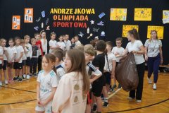 Na zdjęciu widać grupę Uczniów zgromadzonych w sali gimnastycznej podczas wydarzenia sportowego. W tle znajduje się dekoracja z napisem „Andrzejkowa Olimpiada Sportowa” oraz kolorowe plakaty i rysunki. Dzieci ubrane są w stroje sportowe i stoją w grupach, czekając na rozpoczęcie konkurencji. Obok nich są Nauczyciele, którzy pomagają w organizacji wydarzenia. 