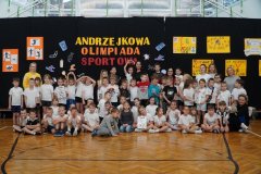 Na zdjęciu widać dużą grupę Dzieci oraz kilku Nauczycieli pozujących do wspólnego zdjęcia w sali gimnastycznej. W tle znajduje się czarna dekoracja z napisem „Andrzejkowa Olimpiada Sportowa” oraz kolorowe plakaty i rysunki przygotowane przez Uczniów. Uczestnicy stoją i siedzą w kilku rzędach, Dzieci mają stroje sportowe. Zdjęcie jest pamiątkową fotografią wszystkich Uczestników wydarzenia.