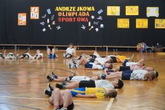 Na zdjęciu widać Dzieci leżące na podłodze w sali gimnastycznej. Są one ubrane w stroje sportowe. Uczniowie wykonują ćwiczenie relaksacyjne po zakończonych zmaganiach sportowych. W tle znajduje się dekoracja z napisem „Andrzejkowa Olimpiada Sportowa” oraz kolorowe plakaty. 