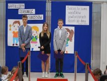 Troje uczniów klasy 8, prowadzących część artystyczną apelu, na tle granatowej ścianki udekorowanej motywem sukcesu.
