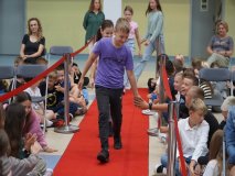 Grupa uśmiechniętych stypendystów z klas 5-6 podążająca po czerwonym dywanie wsród wiwatującej publiczności.