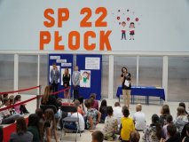 Prezentacja pedagogaga szkolnego, Pani Marty Zawidzkiej, na tle występujących w apelu i ścianki dekoracyjnej.