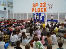 Prezentacja pedagoga specjalnego, Pani Alicji Wojda, na tle występujących w apelu i ścianki dekoracyjnej.