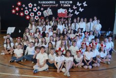 Członkowie chóru, recytatorzy i lektorzy na tle dekoracji, która przedstawia mapę Polski oraz księgę symbolizującą egzemplarz Konstytucji 3 Maja otoczone biało- czerwonymi kwiatami i motylami.