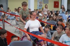 Dziewczynka na wózku inwalidzkim uśmiecha się, przejeżdżając po czerwonym dywanie (pomaga jej kolega) wśród grupy dzieci siedzących na krzesłach i podłodze. W tle widać nauczycielki i uczniów obserwujących wydarzenie.