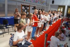 Grupa uczniów stoi na czerwonym dywanie w przestronnej sali, trzymając dyplomy. Obok nich stoi nauczycielka z mikrofonem. W tle widać tablicę z napisem „Starania, Upór, Kreatywność, Ciekawość” oraz siedzących uczniów oglądających uroczystość.