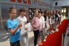 Grupa uczniów stoi na czerwonym dywanie w przestronnej sali, trzymając dyplomy. W tle widać tablicę z napisem „Starania, Upór, Kreatywność, Ciekawość”.