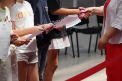 Nauczycielka w czerwonej sukience i białej koszuli wręcza uczennicy dyplom, podając jej rękę. W tle stoją inni uczniowie czekający na odbiór swoich dokumentów