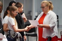 Nauczycielka w czerwonej sukience i białej koszuli wręcza uczennicy dyplom, podając jej rękę. W tle stoją inni uczniowie czekający na odbiór swoich dokumentów