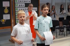 Dwoje uczniów z listami gratulacyjnymi w rękach na czerwonym dywanie w przestronnej sali, w tle klaszcząca Pani Dyrektor oraz tablice z dekoracja w tematyce sukcesu