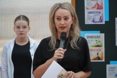 Kobieta przemawia przez mikrofon, a w ręku trzyma dokumenty. Na drugim planie znajduje się ubrana na galowo uczennica, która stoi obok tablicy z dekoracją na temat osiągania sukcesów.