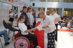 W przestronnej sali na czerwonym dywanie stoi grupa stypendystów, na pierwszym planie uczennica na wózku inwalidzkim, której list gratulacyjny wręcza Pani Dyrektor. Wszyscy są ubrani elegancko. W tle po prawej stronie publiczność złożona z uczniów i nauczycieli, a po lewej przedstawicielki Samorządu Uczniowskiego oraz tablica z tematyczną dekoracją.