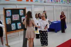 Na czerwonym dywanie stoi jedna z uczennic, a przy niej dwie kobiety, jedna z nich gratuluje dziewczynce sukcesu w konkursie. W tle inna nauczycielka i występujące przedstawicielki Samorządu Uczniowskiego. Po prawej stronie tablica z dekoracją na temat „Klucze do Twojego Sukcesu”.