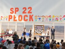 Na tle dużego napisu „SP 22 PŁOCK” oraz rysunków uśmiechniętych dzieci odbywa się przedstawienie. Na scenie znajduje się kilkoro uczniów bierących udział w inscenizacji. Widoczna jest tablica z napisem „KLUCZE DO SUKCESU”, na której znajdują się różne hasła. Część uczniów siedzi przy stoliku, a jedna z uczennic trzyma mikrofon i przemawia. Przed sceną na podłodze siedzi duża grupa dzieci, które uważnie obserwują występ. Po lewej stronie widać Panie Dyrektorki siedzące na krzesłach.