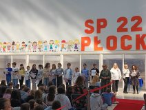 Grupa nagrodzonych stypendystów SP 22 w Płocku stoi na czerwonym dywanie podczas uroczystości, odbierając wyróżnienia, obok uśmiechnięta Pani Dyrektor.