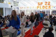 Uczniowie SP 22 w Płocku idą po czerwonym dywanie podczas Apelu Sukcesu, a publiczność uważnie obserwuje. W tle widoczny jest napis szkoły oraz dekoracje.