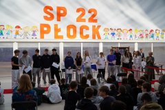 Grupa nagrodzonych stypendystów SP 22 w Płocku stoi na czerwonym dywanie, publiczność złożona z innych uczniów przygląda się z uważnością, obok Pani Dyrektor bije brawo.