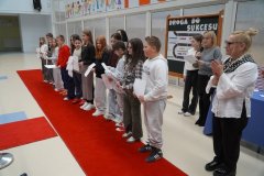 W przestoronnej sali z dużym napisem w tle: "SP 22 PŁOCK", na czerwonym dywanie stoi rząd uczniów z listami gratulacyjnymi w dłoniach, obok stoją dwie kobiety i dziewczyna- biją brawo.