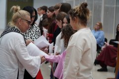 Kilkoro uśmiechniętych uczniow ustawionych w szeregu, dwie kobiety wręczają im listy gratulacyjne oraz ściskają dłoń podczas gratulacji.