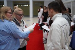 Kilkoro uśmiechniętych uczniow ustawionych w szeregu na czerwonym dywanie, dwie kobiety wręczają im listy gratulacyjne oraz ściskają dłoń podczas gratulacji.