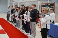 W przestoronnej sali na czerwonym dywanie stoi rząd uczniów z listami gratulacyjnymi w dłoniach, obok stoi kobieta i bije brawo.