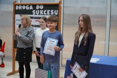 W przestronnej sali przed dekoracyjną tablicą z hasłem "Droga do sukcesu" stoi dwoje radosnych uczniów, w dłoniach trzymają dyplomy oraz torebli ozdobne z nagrodami konkursowymi, nieopodal stoi kobieta przemawiająca przez mikrofon oraz inna uczennica.