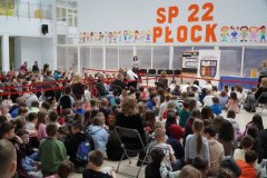 Duża sala szkolna, na któej zgromadzonych jest wielu uczniów i nauczycieli. W tle: stół z granatowym obrusem, baner z logo szkoły oraz tablica z dekoracją o sukcesie, a także duży pomarańczowy napis na ścianie: "SP 22 W PŁOCKU", po obu stronach kolorowe ilustracje dzieci.