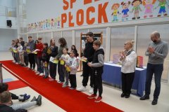 W przestoronnej sali z dużym napisem w tle: "SP 22 PŁOCK", na czerwonym dywanie stoi rząd uczniów z listami gratulacyjnymi w dłoniach, obok stoi mężczyazna i kobieta, którzy biją brawo.