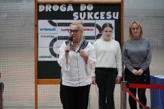 Kobieta trzyma mikrofon i przemawia z uśmiechem, w tyle stoi kobieta i uczennica, obok znajduje się tablica z dekoracją dotyczącą sukcesu- ilustracja zawiłej drogi z wieloma napisami, np. wytrwałość, dyscyplina, uczciwość.