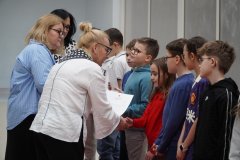 Kilkoro uśmiechniętych uczniow ustawionych w szeregu, dwie kobiety wręczają im listy gratulacyjne oraz ściskają dłoń podczas gratulacji.