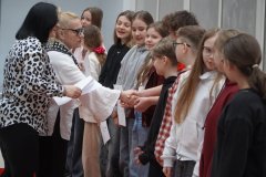 Kilkoro uśmiechniętych uczniow ustawionych w szeregu, dwie kobiety wręczają im listy gratulacyjne oraz ściskają dłoń podczas gratulacji.