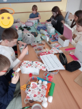 1 Grupa uczniów siedzących wokół dużego stołu w sali lekcyjnej. Dzieci są pochłonięte pracą plastyczną – ozdabiają białe kartonowe pudełka, które przekształcają w apteczki pierwszej pomocy emocjonalnej. Na stole rozłożone są różnorodne materiały: kolorowe papiery (w tym serwetki w serduszka), kleje, nożyczki, taśmy dekoracyjne, flamastry, wstążki, wycinanki z pianki oraz pudełka z kredkami i pisakami. Każde dziecko pracuje indywidualnie, według własnego pomysłu, ale panuje atmosfera skupienia i zaangażowania. 