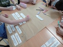 Foto_12 Uczniowie pracują w sali lekcyjnej wokół wspólnego stołu. Tworzą zespołowo plakat dotyczący radzenia sobie z emocjami. Na brązowym arkuszu papieru układają i przyklejają przygotowane wcześniej zdania z poradami oraz kolorowo ozdabiają tytuł pracy PRZYJAŻŃ.
