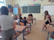 Foto_16 Grupa uczniów w sali lekcyjnej podczas zabawy integracyjnej. Jeden z uczestników trzyma ręce w pudełku , w którym znajduje się jakiś przedmiot i próbuje, nie widząc go, określić co to jest. Wokół inni uczestnicy zajęć kibicują chłopcu. W tle zielona tablica z baiłymi napisami: "Skarby", "Optymizm", "Przyjaźń", "Kreatywność.