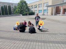 Foto_17 Plac przed szkołą, grupa uczestników zajęć podczas zabawy integracyjnej. Wszyscy skupieni wokół siebie w kręgu, większość z nich siedzi, jedna dziewczynka wskazuje na innego uczestnika.