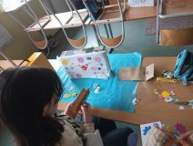 Foto_3 Uczestniczka zajęć ozdabia swoją apteczkę w białą bibułę i kolorowe pankowe kwiatki i kształty, w tle otoczenie sali lekcyjnej: ławki i krzesła.