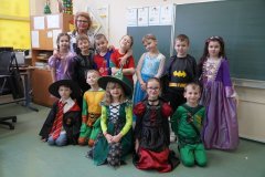 Przebrani za księżniczki, czarodziejów i superbohaterów uczniowie klasy 1D, wraz z wychowawcą, pozują do grupowego zdjęcia na tle tablicy szkolnej.