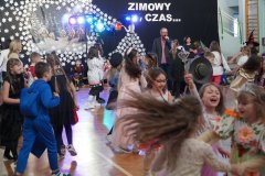 Uczniowie tańczą na sali gimnastycznej. W tle znajduje się nauczyciel prowadzący zabawę, reflektory oraz zimowa dekoracja.