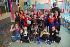 Zdjęcie grupowe Uczniów klasy 1f. Na tle karnawałowej dekoracji stoją Dzieci przebrane m.in. za superbohaterów, syrenkę i księżniczki.