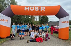 Reprezentacja Szkoły Podstawowej nr 22 w Płocku na Powiatowych Sztafetowych Biegach Przełajowych  - grupa dziewcząt i chłopców w niebieskich strojach sportowych pozująca pod pomarańczowo – białą – czarną bramą MOSiR Płock