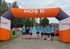Reprezentacja Szkoły Podstawowej nr 22 w Płocku na Powiatowych Sztafetowych Biegach Przełajowych  - grupa chłopców  w niebieskich sportowych koszulkach z numerami stojąca pod pomarańczowo – białą – czarną bramą MOSiR Płock