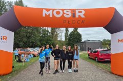 Reprezentacja Szkoły Podstawowej nr 22 w Płocku na Powiatowych Sztafetowych Biegach Przełajowych  - grupa dziewcząt  w niebieskich sportowych koszulkach z numerami stojąca pod pomarańczowo – białą – czarną bramą MOSiR Płock