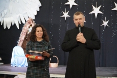 Wicedyrektor Szkoły- p. A. Gawlińska oraz ksiądz P. Szulc, stojąc na tle dekoracji, ogłaszają wyniki konkursu na najpiękniejszą szopkę bożonarodzeniową.