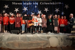 Chłopiec w czapce Mikołaja gra na gitarze w tle chór w świątecznych strojach.