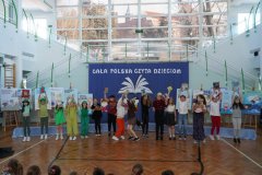 Na tle dekoracji stoi grupa dzieci z podniesionymi do góry rękami. W rękach trzymają egzemplarze książek z wierszami Juliana Tuwima.