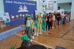 Na tle dekoracji stoją dzieci ustawione gęsięgo w kolorowych strojach  do prezentacji wiersza ,,Rzepka”. Przed pierwszym dzieckiem leży rekwizyt wykonany z bibuły /Rzepka.