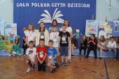 Na tle dekoracji stoi grupa dzieci z 2 nauczycielami. Dzieci na szyjach mają zawieszone medale za czytelnictwo.