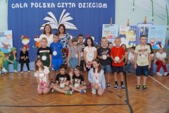 Na tle dekoracji stoi grupa dzieci z 2 nauczycielami. Dzieci na szyjach mają zawieszone medale za czytelnictwo.